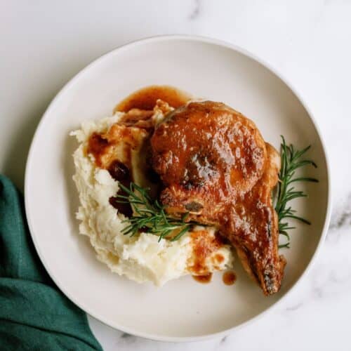 A plate with mashed potatoes, a pork chop topped with brown gravy, and a sprig of rosemary, on a white surface next to a green napkin.
