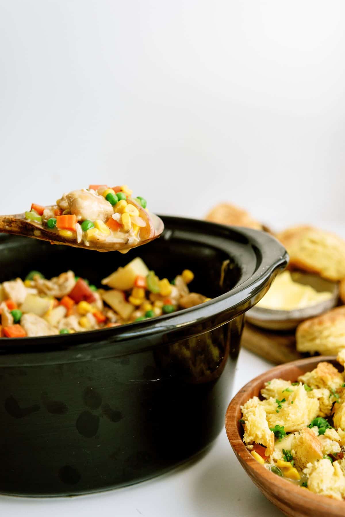 A scoop of Slow Cooker Chicken Pot Pie on a wooden spoon above the slow cooker. A bowl of Slow Cooker Chicken Pot Pie and a plate of biscuits