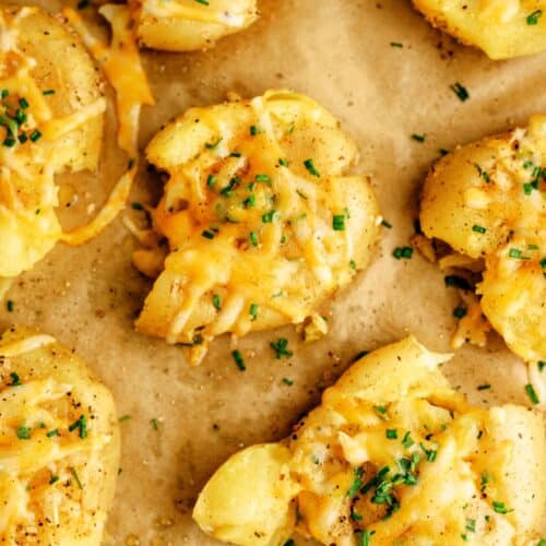 Baked smashed potatoes topped with melted cheese and chopped chives on parchment paper.