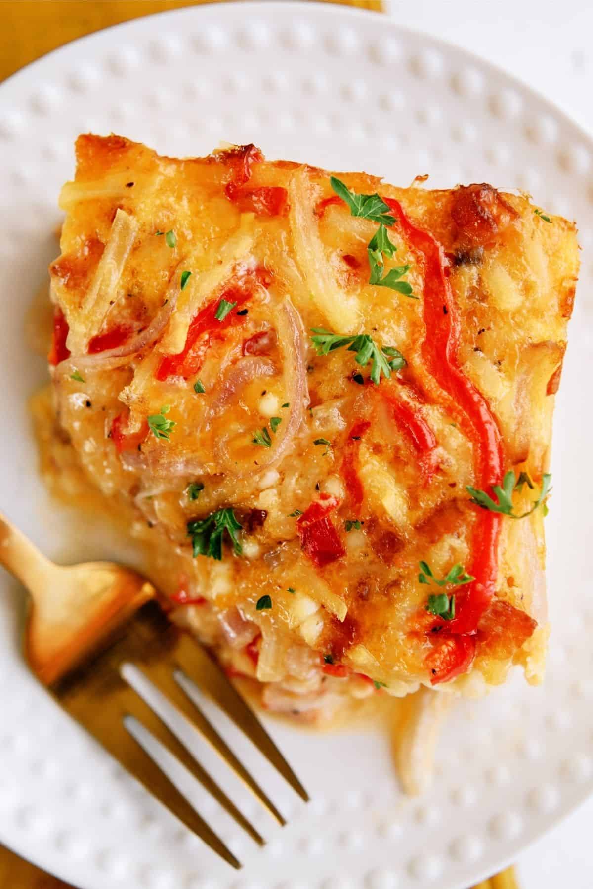 A piece of cheesy casserole with red peppers and herbs on a white plate, accompanied by a gold fork.