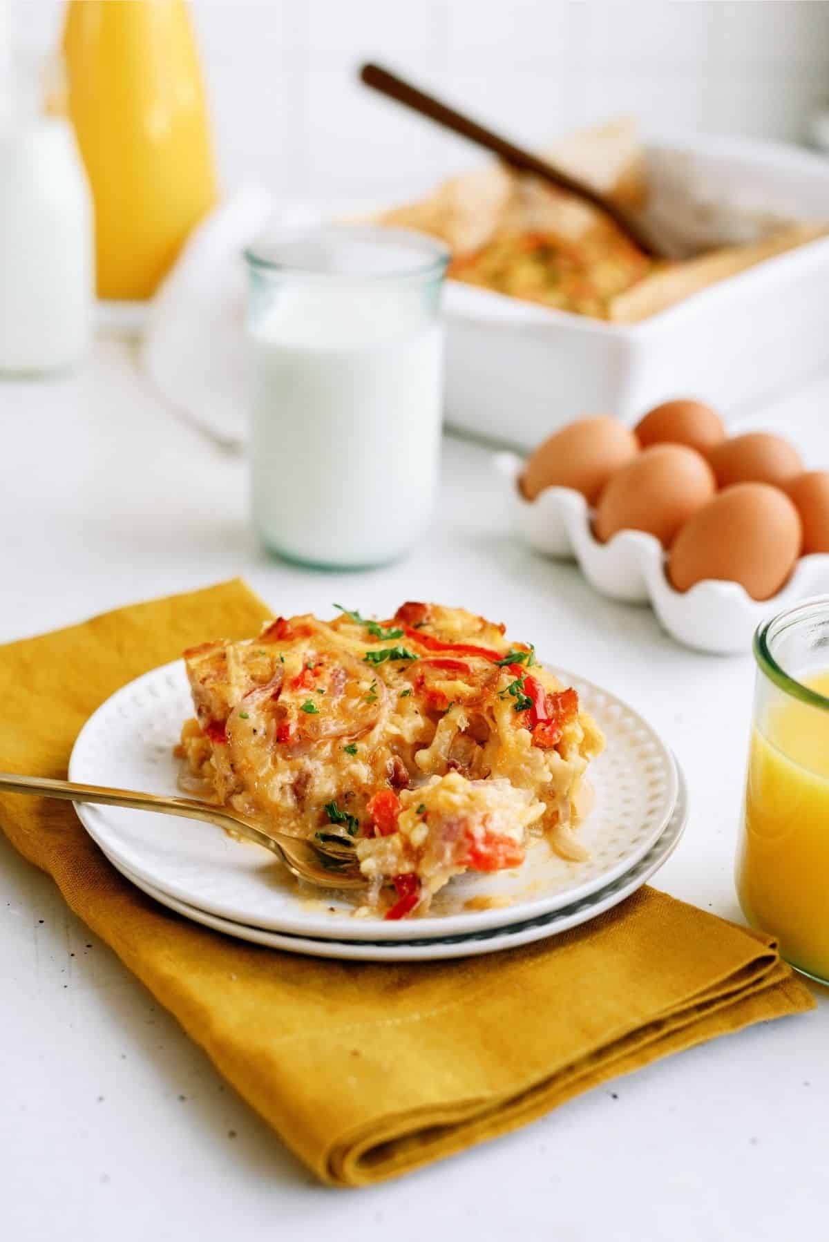 A slice of casserole on a plate with a fork, surrounded by glasses of milk and orange juice, a bowl of brown eggs, and a casserole dish on a yellow cloth.