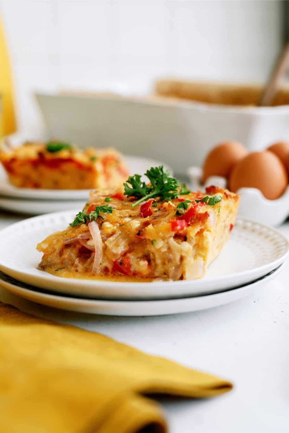 A slice of Amish breakfast casserole with parsley on a white plate, with whole eggs and a baking dish in the background.