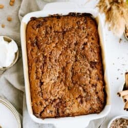 A baked caramel chip cake in a white rectangular dish, accompanied by a bowl of whipped cream, plates, a cup of milk, cinnamon sticks, and dried flowers.