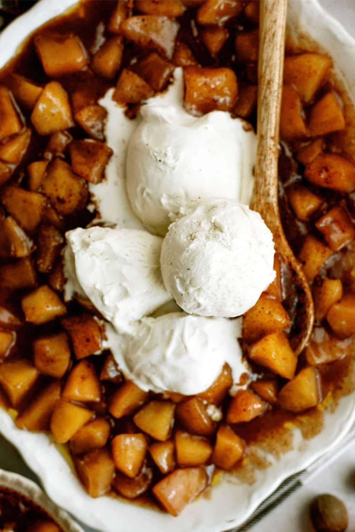 A serving bowl with a wooden spoon filled with Copycat Cracker Barrel Cinnamon Apples and topped with 3 scoops of vanilla ice cream.