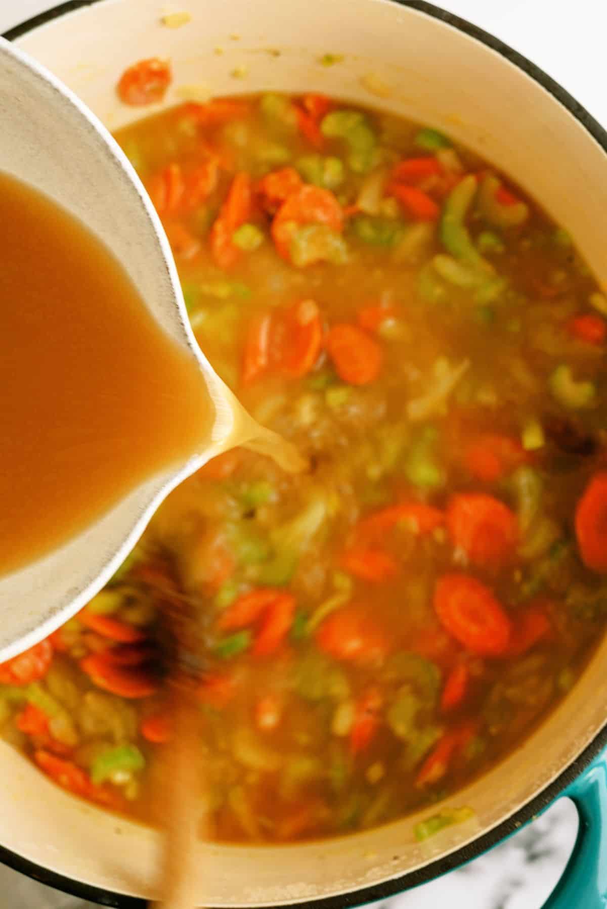 Pouring broth from measuring cup into stock pot full of chopped veggies.
