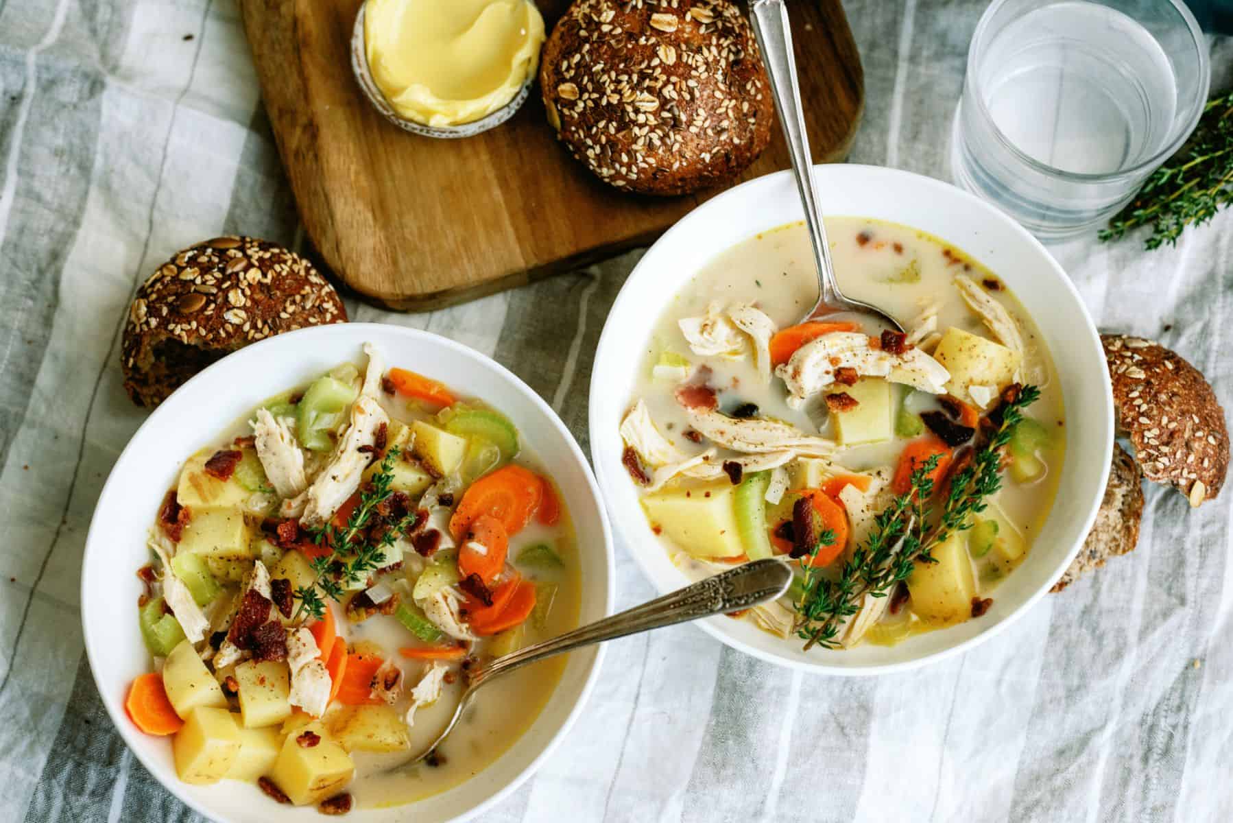 2 bowls of Creamy Chicken and Potato Soup with spoons, rolls on a cutting board with a small dish of butter.