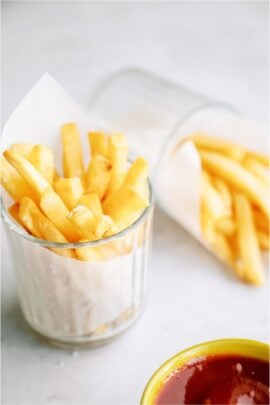 Two small glasses filled with Crispy Air Fryer Frozen French Fries wrapped in a paper towel with a side of ketchup off to the side.