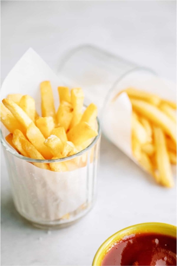 Two small glasses filled with Crispy Air Fryer Frozen French Fries wrapped in a paper towel with a side of ketchup off to the side.