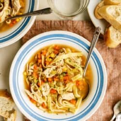 A bowl of chicken noodle soup with vegetables sits on a striped plate, next to a glass of water, sliced bread, and a pink napkin with utensils.