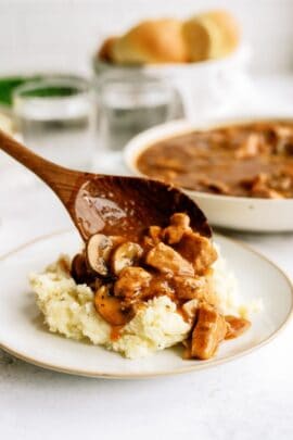 A plate of mashed potatoes with a wooden spoon spooning a serving of Instant Pot Pork Tips and Gravy on top of it.