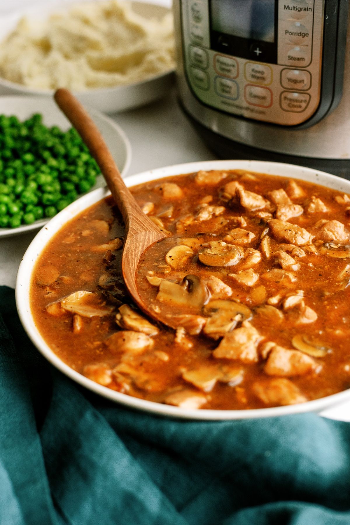 A serving dish full of Instant Pot Pork Tips and Gravy with an Instant Pot in the background and a side of peas