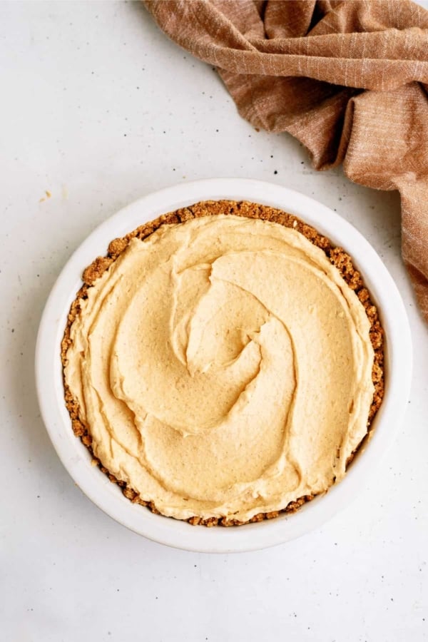 A pie with a graham cracker crust and a smooth, creamy filling is placed on a white surface next to a brown cloth.