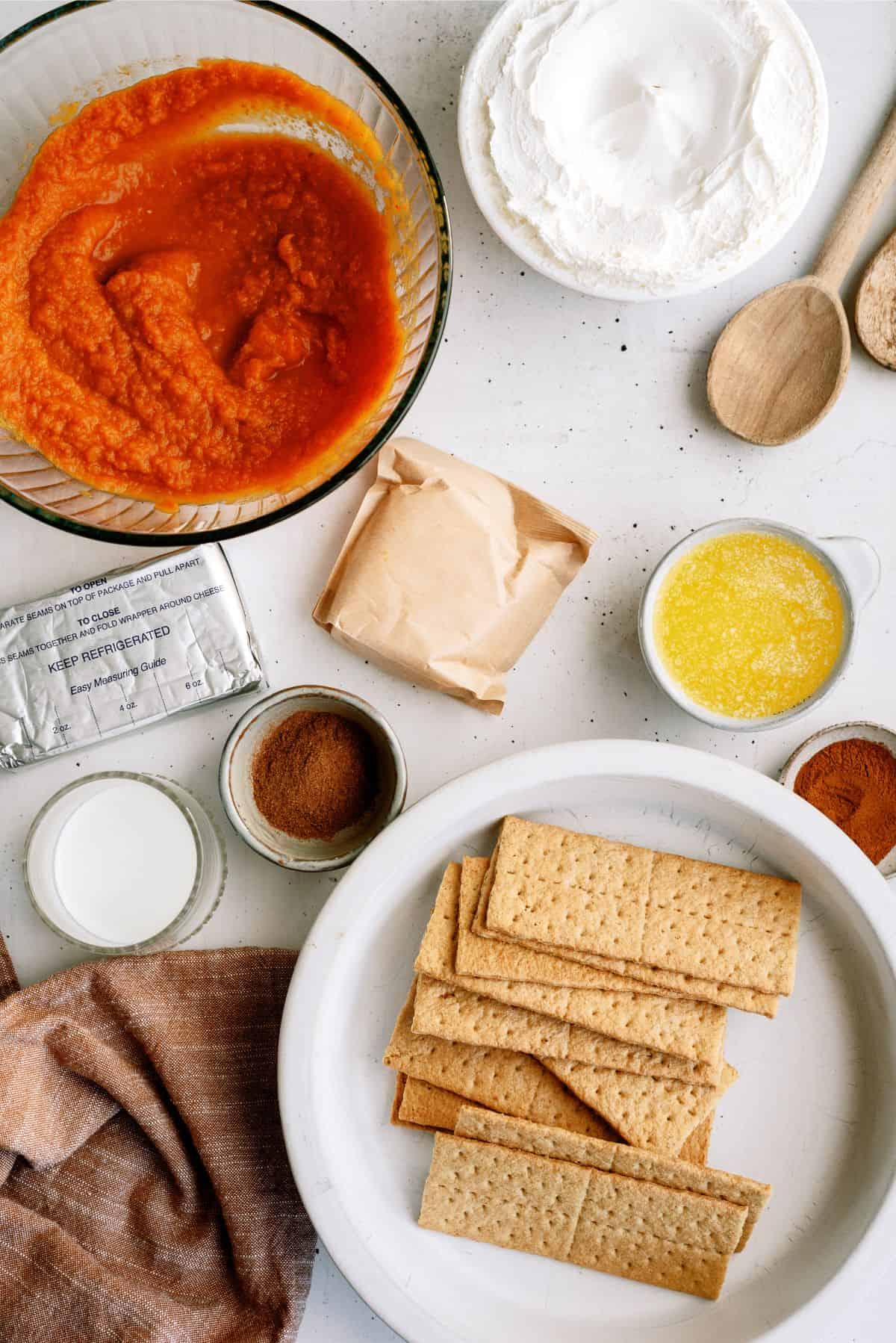 Ingredients for making a pumpkin cheesecake are arranged on a table, including pureed pumpkin, cream cheese, graham crackers, melted butter, spices, and sugar.