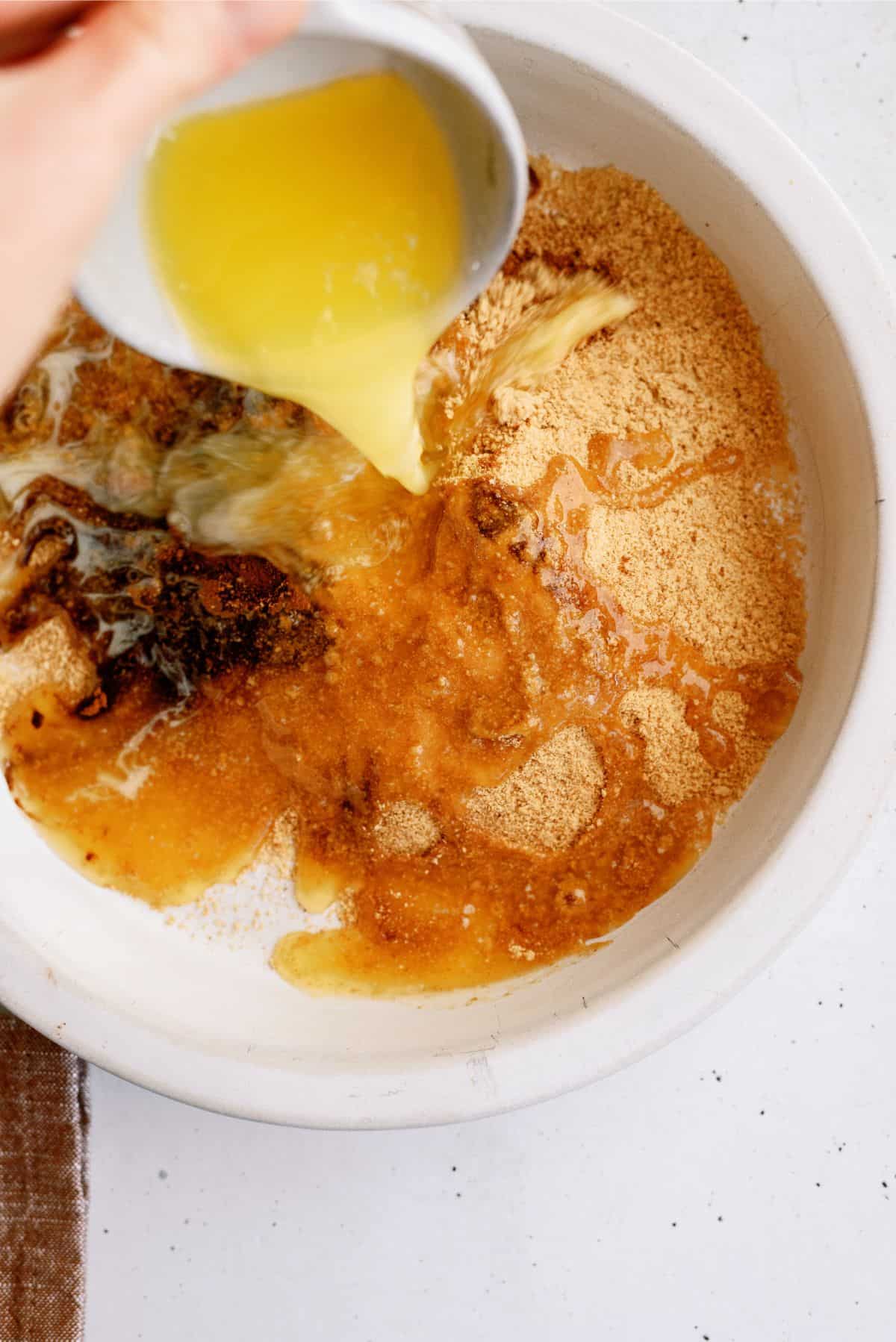 A hand is pouring melted butter from a small white bowl into a mixing bowl containing brown sugar and other ingredients.