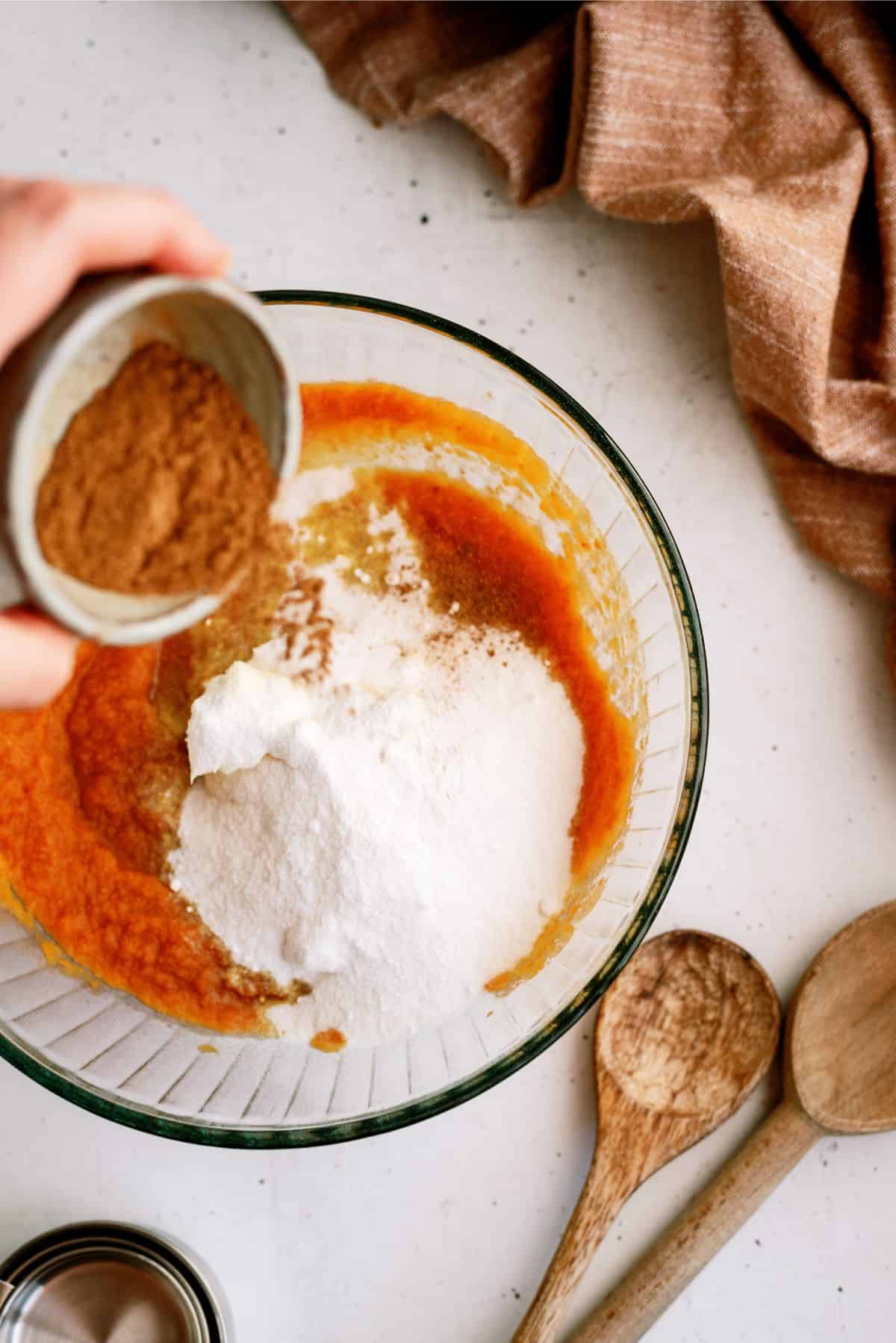 A hand pours a small bowl of cinnamon into a glass mixing bowl containing flour and an orange liquid mixture. Wooden spoons and a brown cloth are nearby on a white surface.