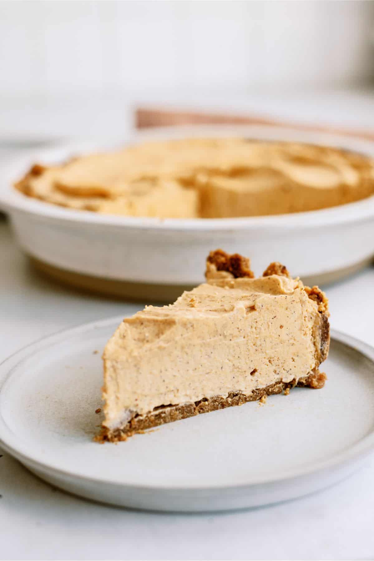 A slice of pumpkin pie served on a white plate with the remaining pie in the background.