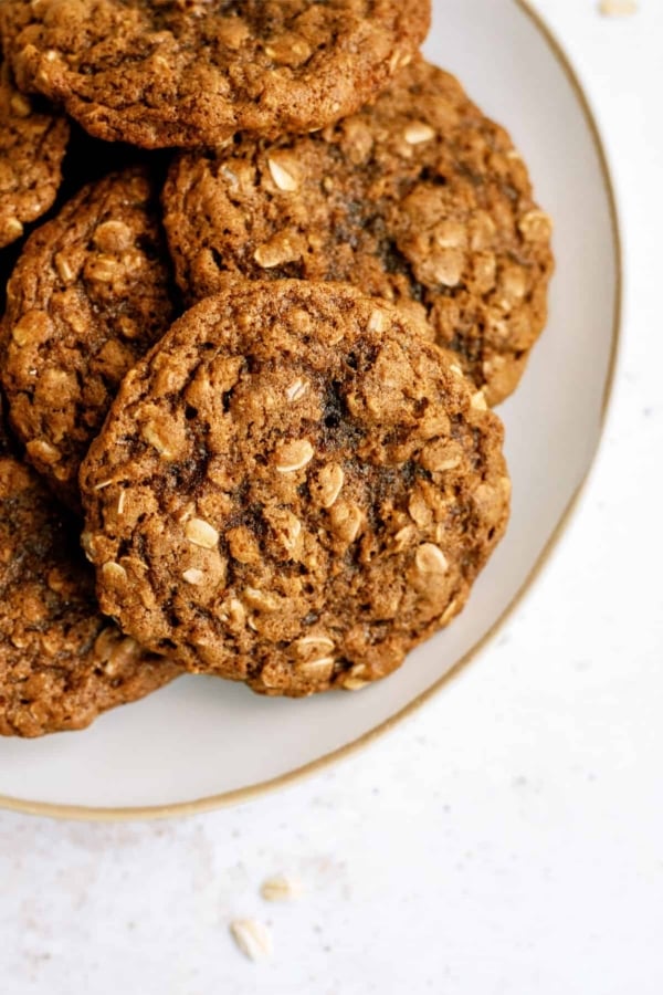 A close-up of a plate with several oatmeal cookies. The cookies have a coarse texture and visible oats. The plate is white with a gold rim and is placed on a white surface.