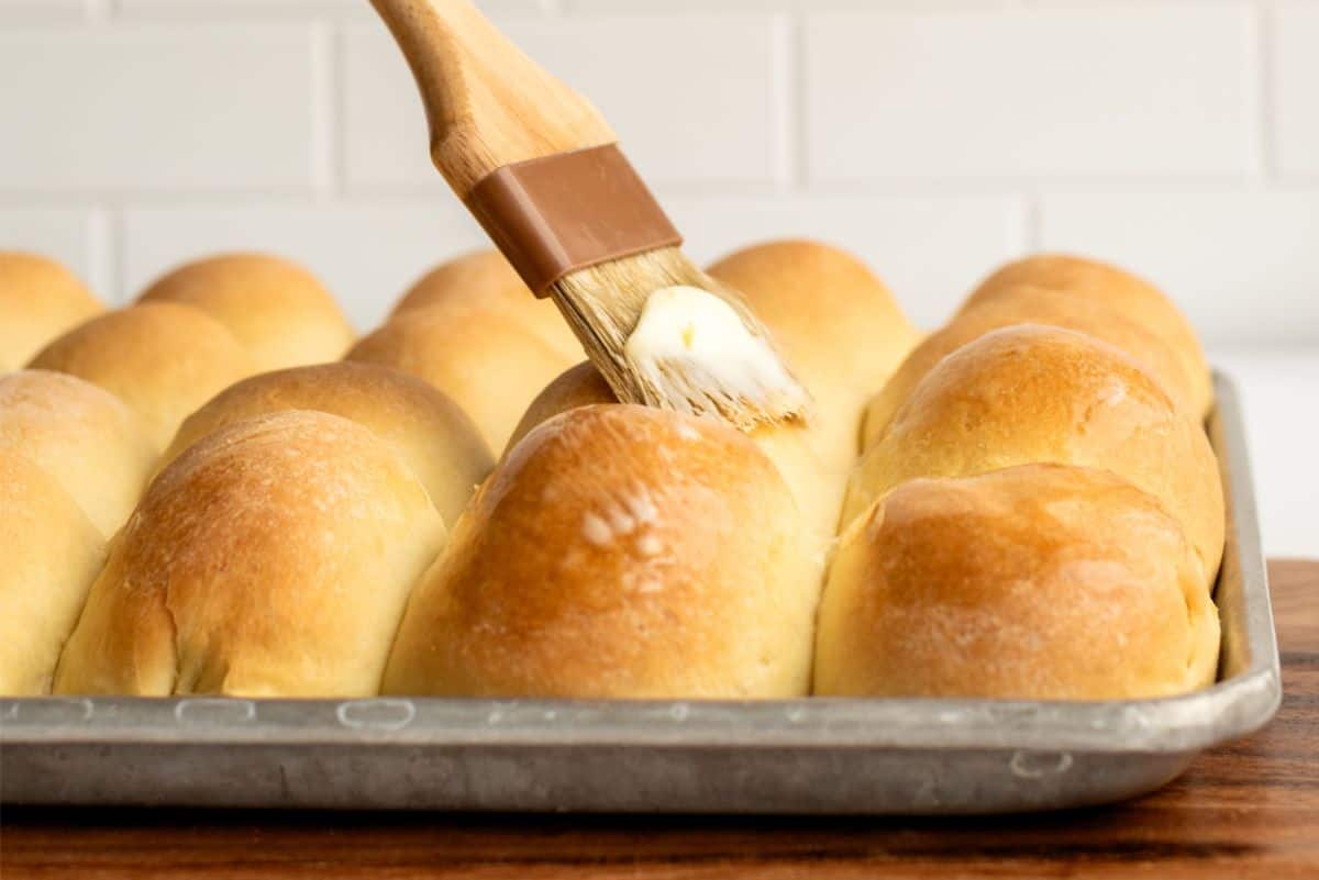 A brush spreads butter over a tray of freshly baked dinner rolls.