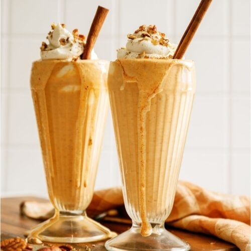 2 glasses of Pumpkin Pie Milkshakes topped with whip cream and cinnamon sticks.