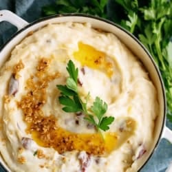 A pot of mashed potatoes with garlic, butter, and parsley garnish, set on a blue cloth with fresh parsley and garlic in the background.