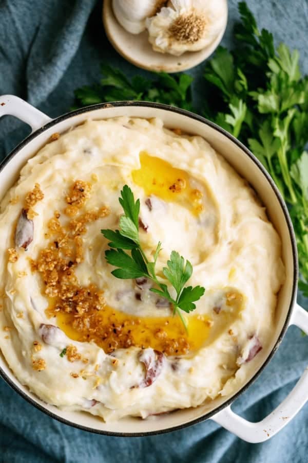 A pot of mashed potatoes with garlic, butter, and parsley garnish, set on a blue cloth with fresh parsley and garlic in the background.
