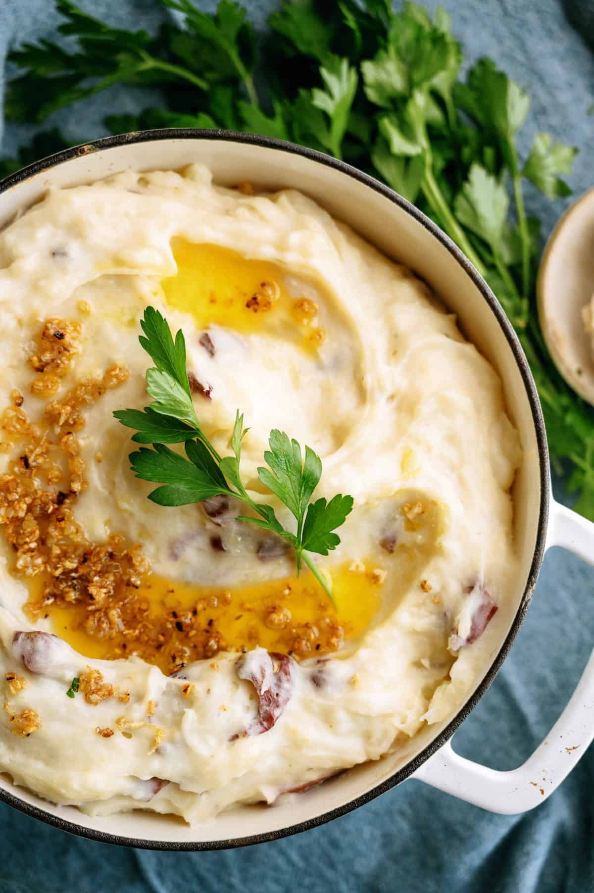 A pot of mashed potatoes with melted butter, crispy garlic bits, and fresh parsley garnish, set on a blue cloth with parsley leaves in the background.
