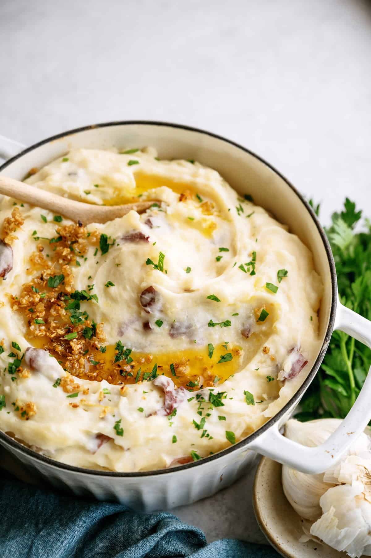 A pot of mashed potatoes with herbs and melted butter on top, garnished with chopped parsley, with a wooden spoon resting inside.