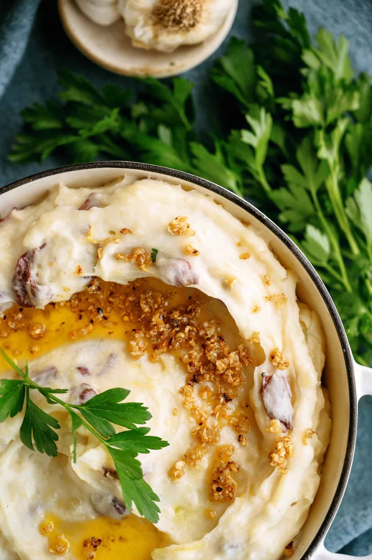 A bowl of mashed potatoes topped with melted butter, crispy garlic bits, and garnished with fresh parsley; fresh parsley leaves are placed nearby.