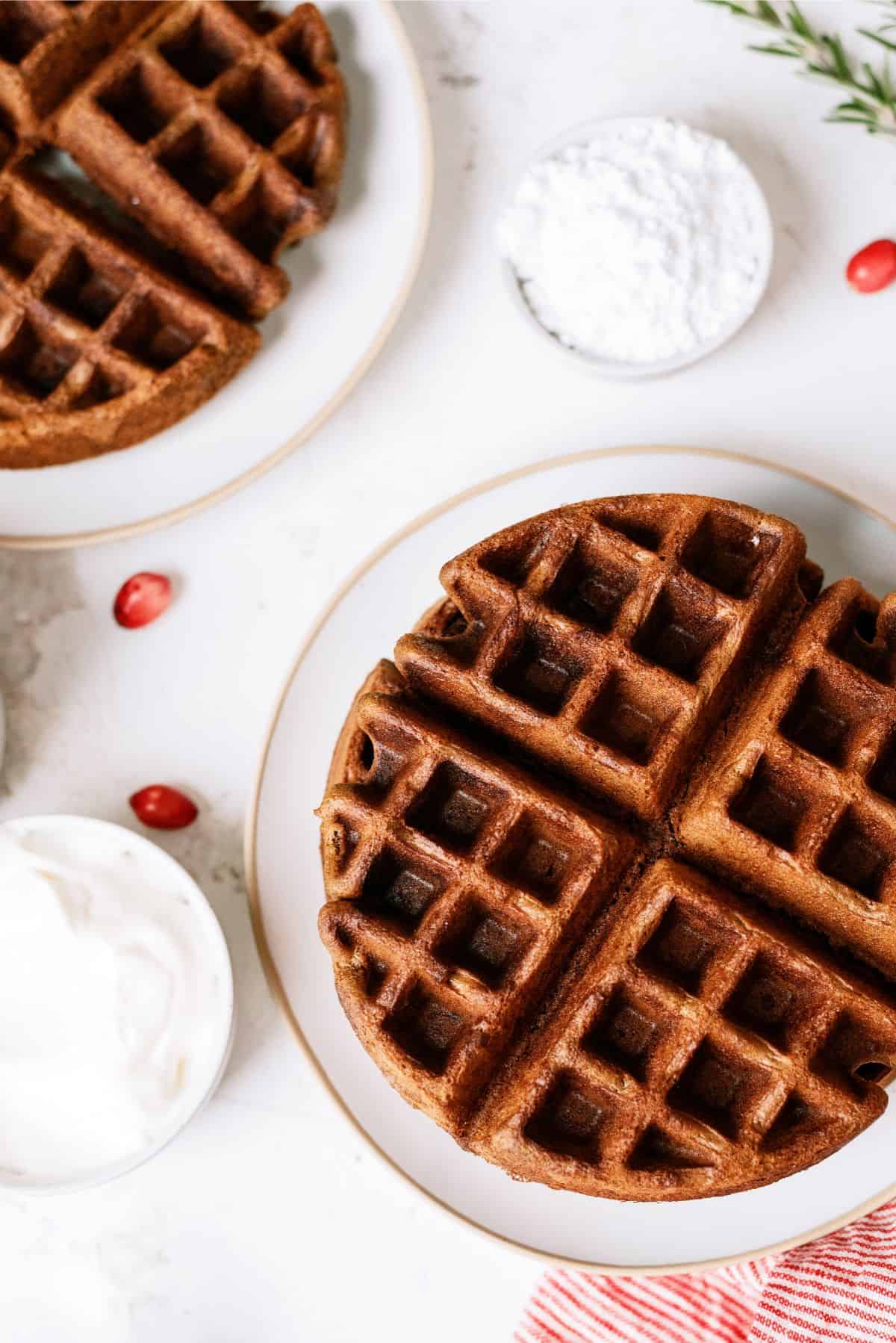 A plate with a whole circular waffle, a bowl of whipped cream, and a bowl of powdered sugar. There are also red berries scattered on the white surface.
