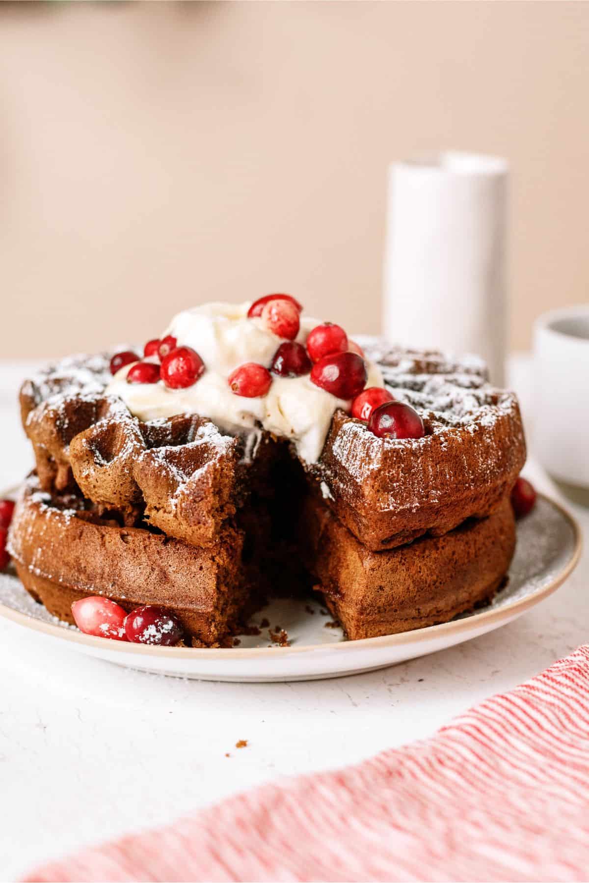 A stack of gingerbread waffles topped with cream and cranberries, partially sliced, on a plate.