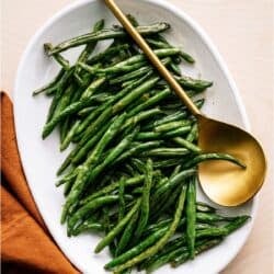 A white oval plate filled with cooked green beans sits next to a gold serving spoon and an orange cloth napkin on a light surface.