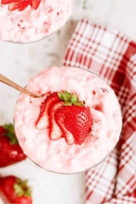 A glass bowl filled with pink strawberry mousse, topped with sliced fresh strawberries, sits on a white surface near a red and white plaid napkin.