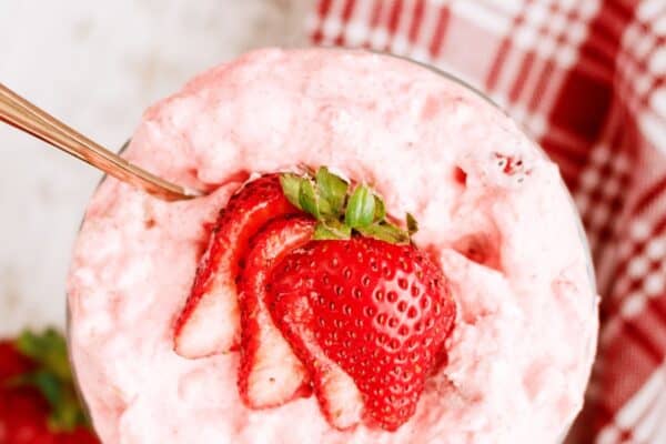 A glass bowl filled with pink strawberry mousse, topped with sliced fresh strawberries, sits on a white surface near a red and white plaid napkin.