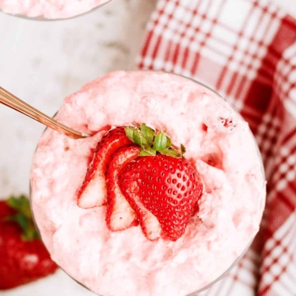 A glass bowl filled with pink strawberry mousse, topped with sliced fresh strawberries, sits on a white surface near a red and white plaid napkin.