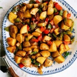 A plate of seasoned roasted potatoes mixed with chopped red and green bell peppers on a floral-patterned dish, with a fork beside it.