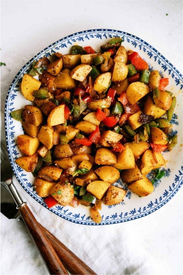 A plate of seasoned roasted potatoes mixed with chopped red and green bell peppers on a floral-patterned dish, with a fork beside it.