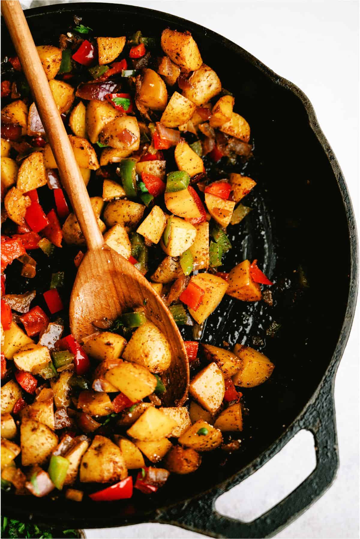 Diced potatoes, bell peppers, and herbs cooking in a black skillet with a wooden spoon.