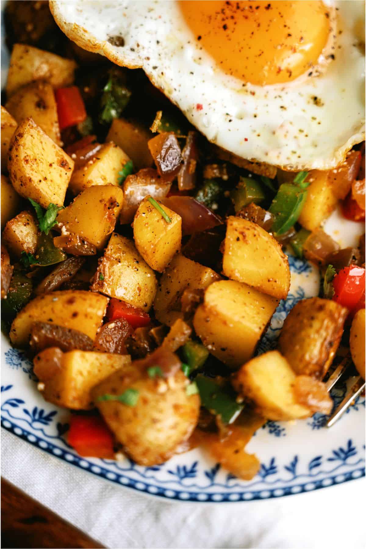 A close-up of a plate with seasoned diced potatoes, vegetables, and a sunny-side-up egg on top, served on a decorative white and blue plate.