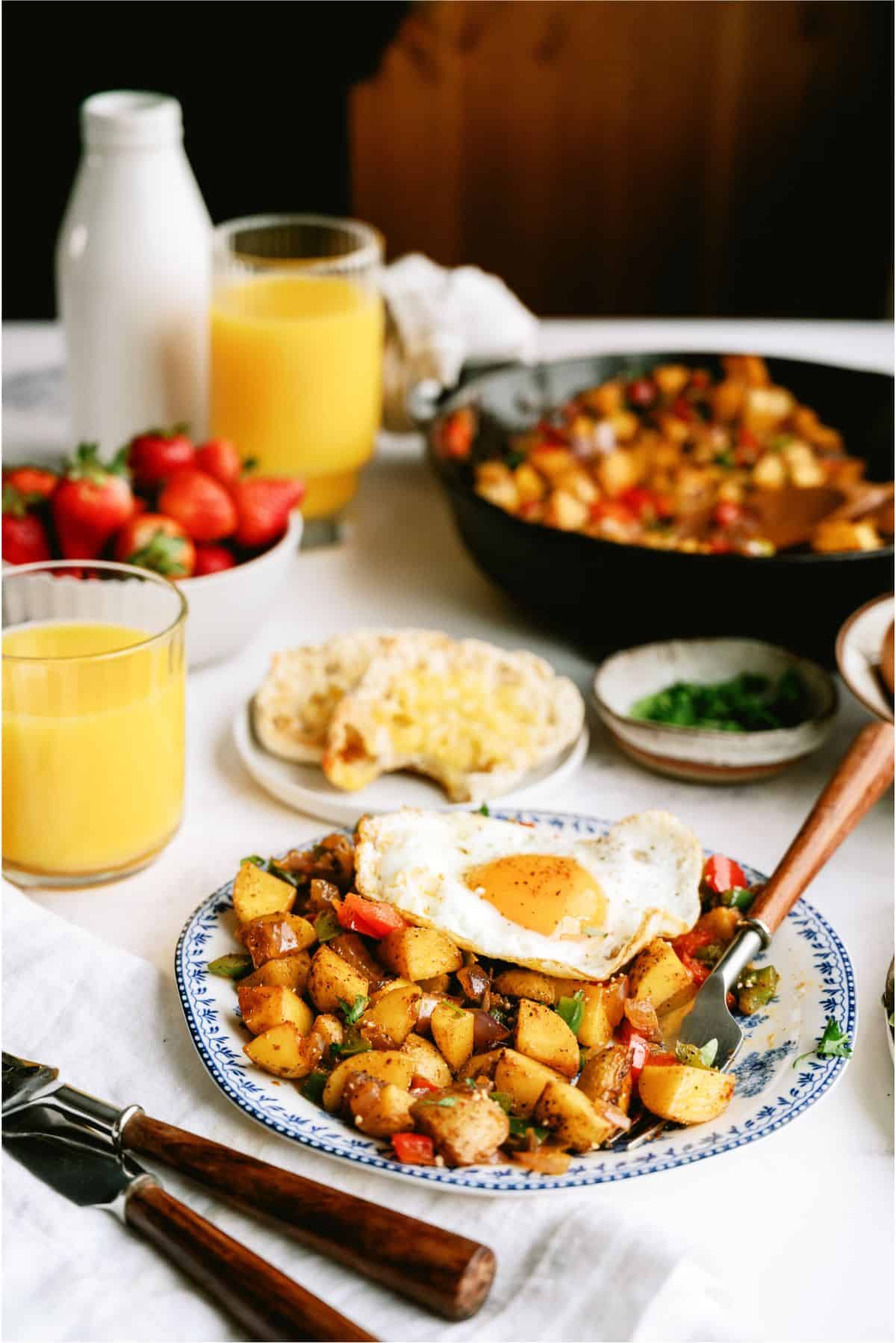 A breakfast table with a plate of fried eggs and seasoned potatoes, a glass of orange juice, strawberries, and various condiments.