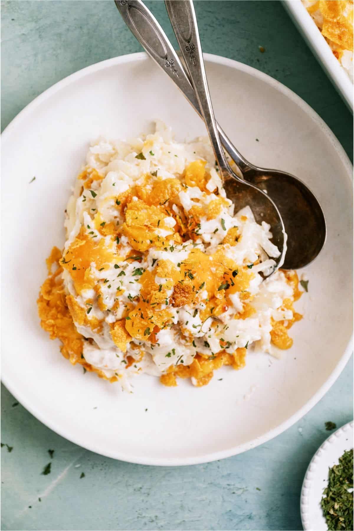 A white plate with a serving of Cheesy Hashbrowns Potato Casserole (Funeral Potatoes) on it and two spoons.