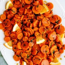 Sliced cooked carrots garnished with chopped herbs and orange wedges on a white serving platter.