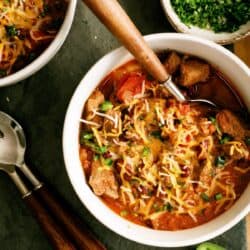 A bowl of chili topped with shredded cheese and chopped herbs, with a spoon inside. Sliced jalapeños, another bowl of chili, and a bowl of herbs are nearby on a dark surface.