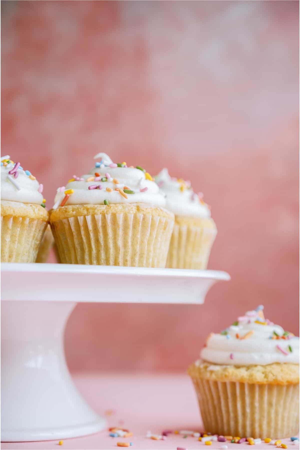 Easy Vanilla Cupcakes on a cake stand with one on the countertop. All topped with frosting and sprinkles.