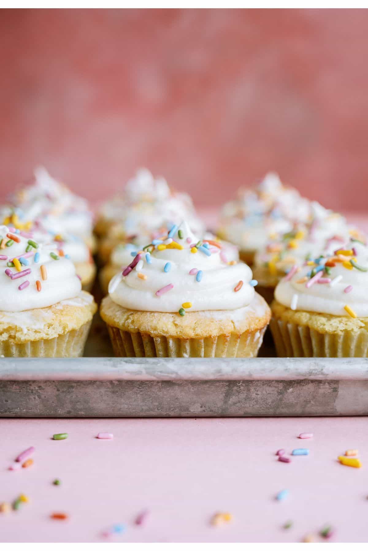 Easy Vanilla Cupcakes topped with frosting and sprinkles lined in rows on a baking sheet.