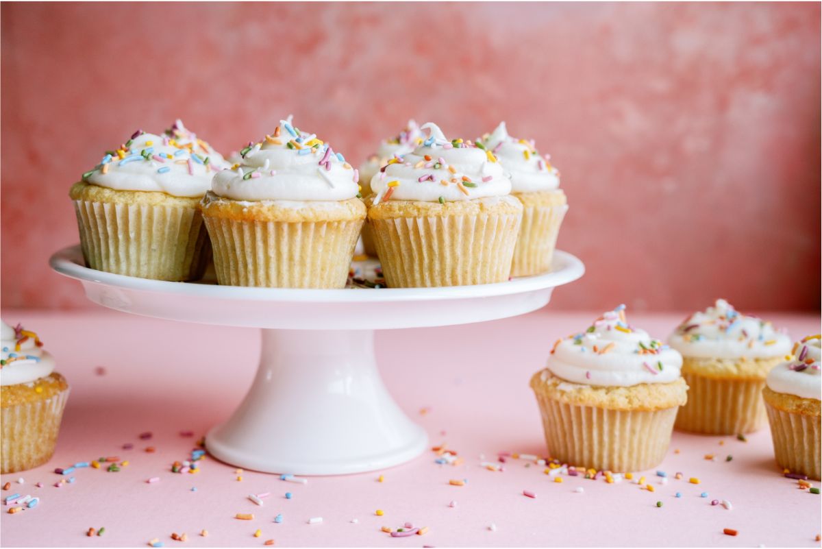 Frosted vanilla cupcakes with sprinkles on a cake stand with some also on the counter