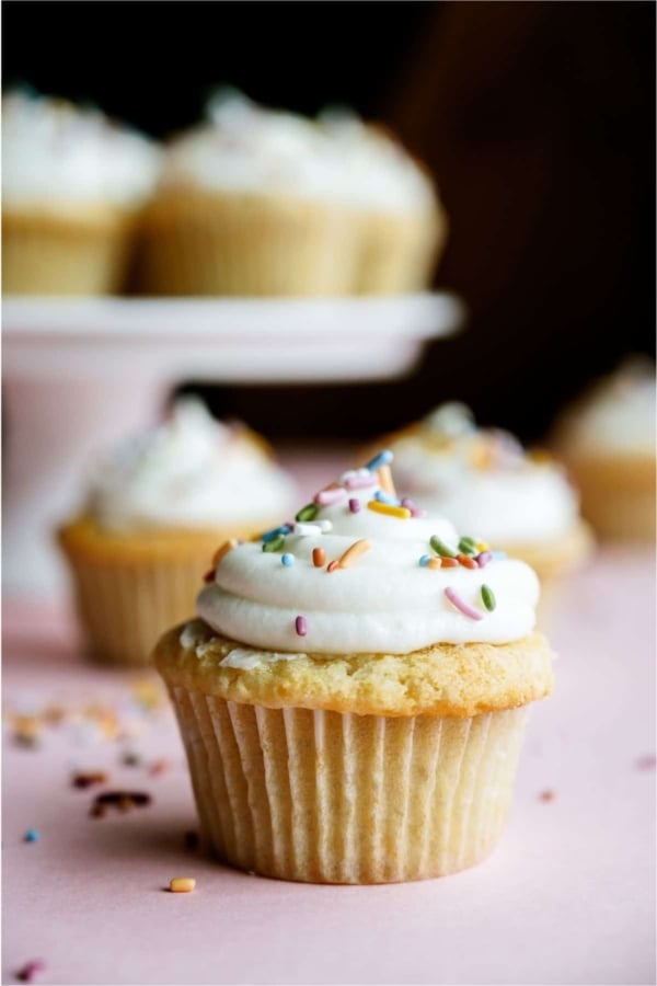 Easy Vanilla Cupcake with frosting and sprinkles. On a pink background. With other cupcakes in the background.