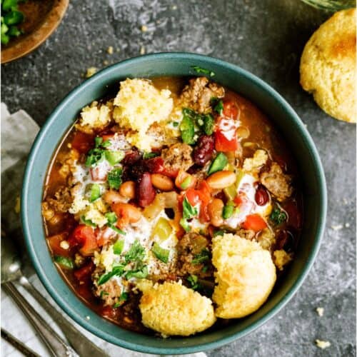 A bowl of chili topped with beans, ground meat, tomatoes, cheese, and cilantro, served with cornbread on the side. Two spoons and a napkin are placed next to the bowl.