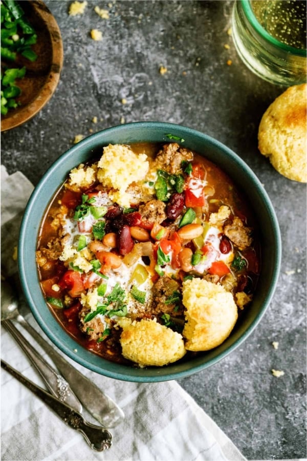 A bowl of chili topped with beans, ground meat, tomatoes, cheese, and cilantro, served with cornbread on the side. Two spoons and a napkin are placed next to the bowl.