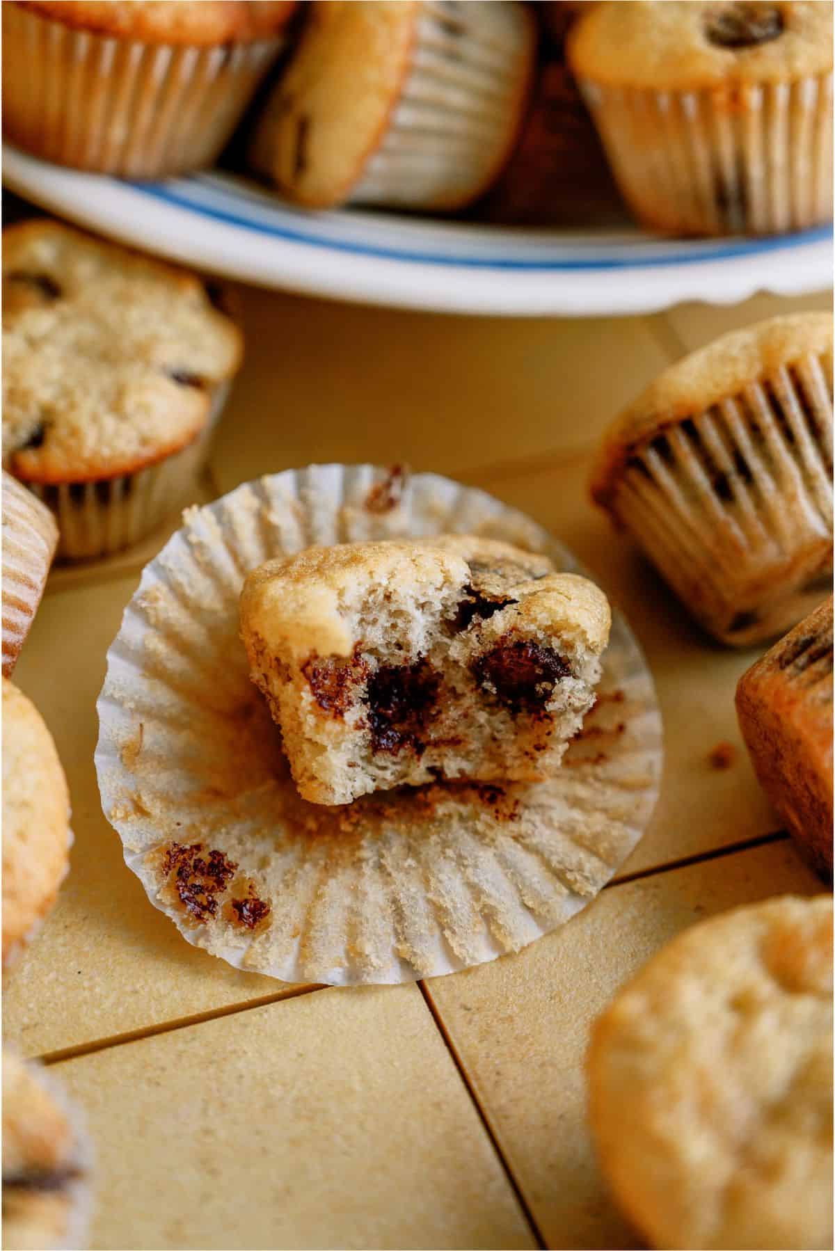 A partially eaten muffin with visible chocolate chips sits on a crinkled paper liner, surrounded by other whole muffins on a plate and the table.