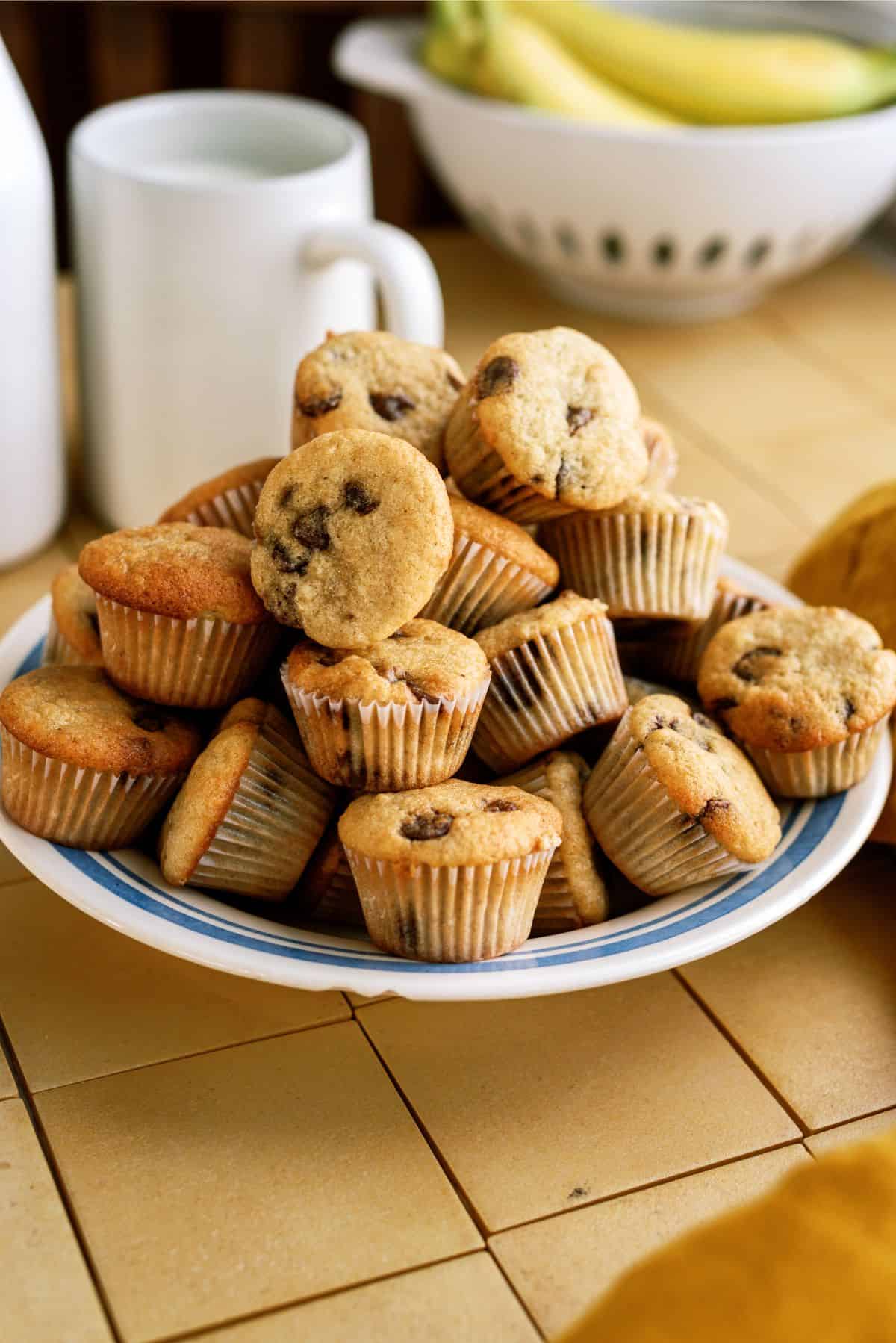 A plate filled with mini muffins, some with chocolate chips, sits on a tiled countertop. A mug, a bottle, and a bowl of bananas are in the background.
